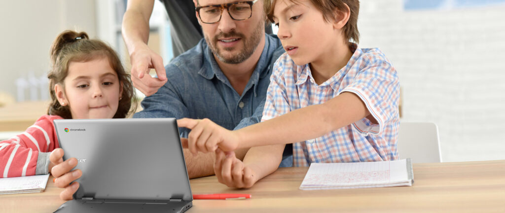 A male adult and two children sitting at a desk looking engagingly at a Chromebook device.